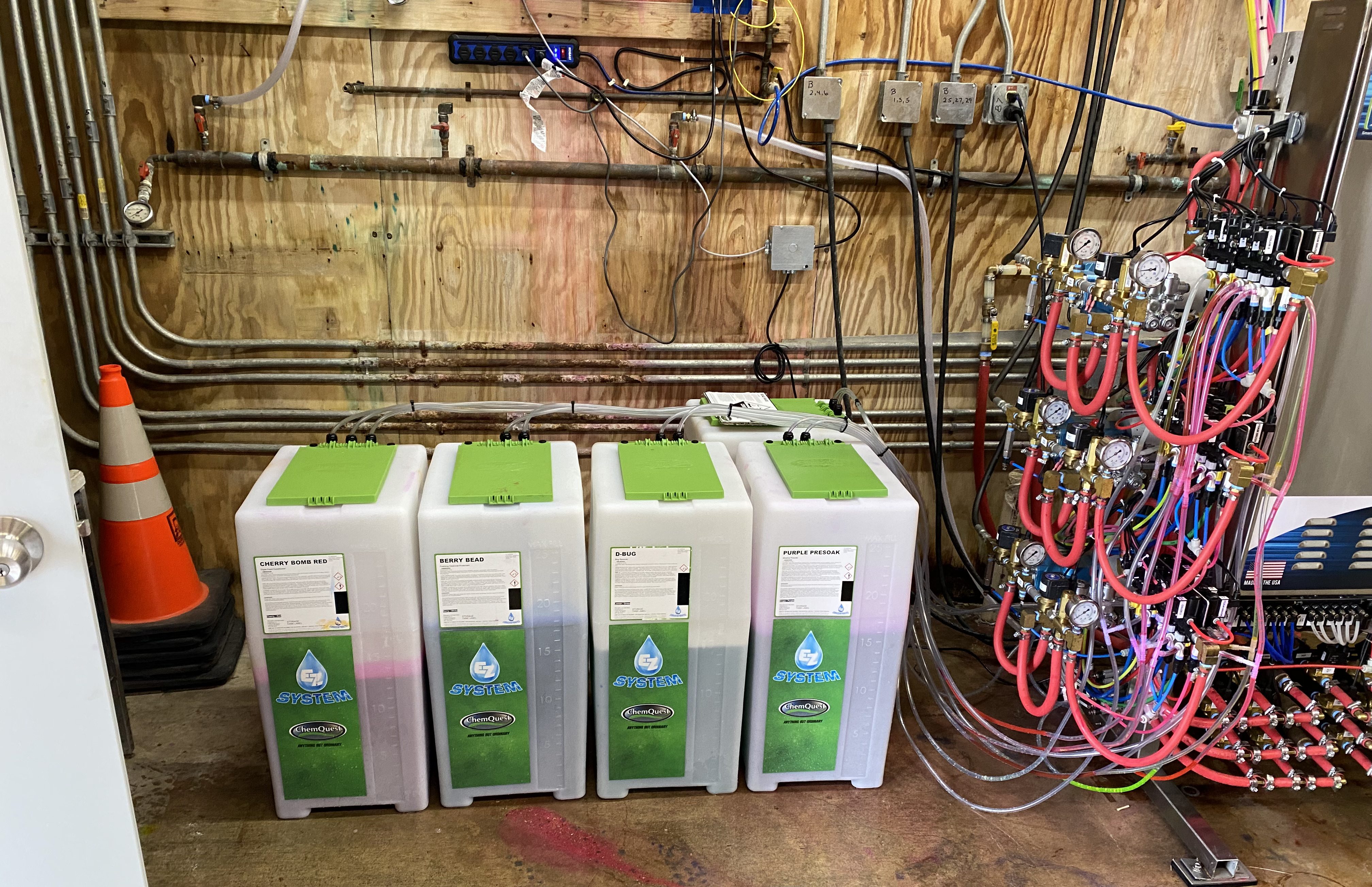 Four EZ System car wash chemical storage tanks lined up against an equipment room wall with chemical lines to the proportioning system