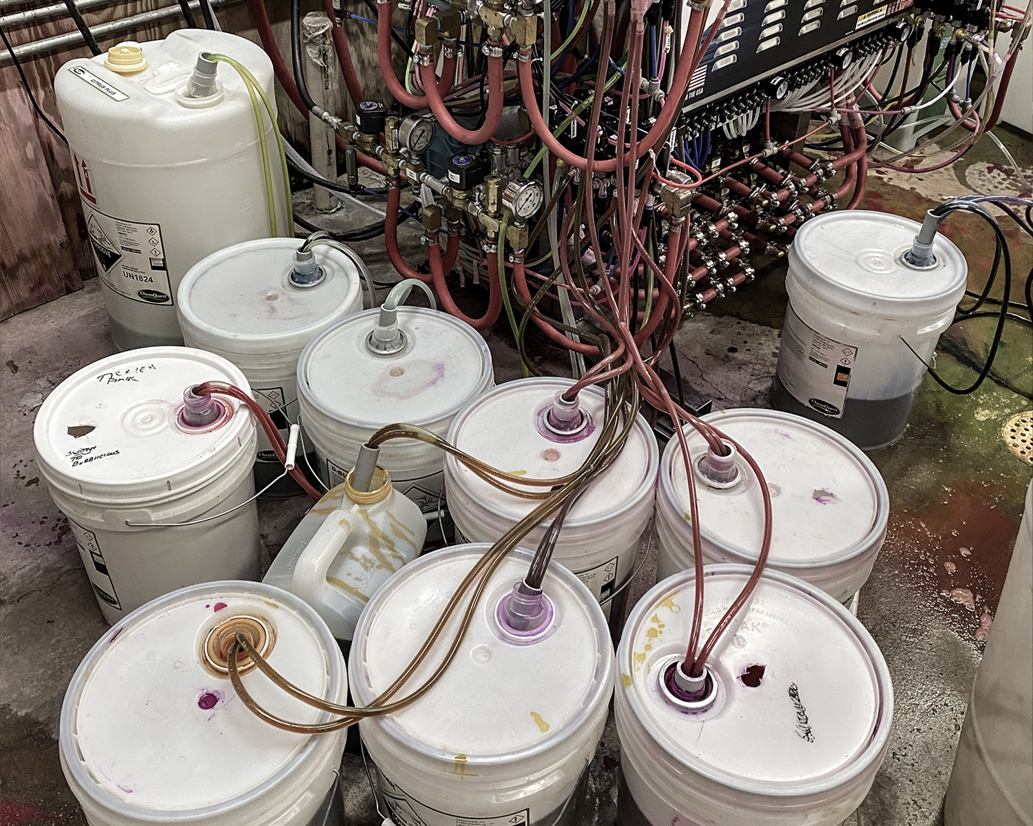 Car wash equipment room crowded with chemical pails, drums and tangled feed lines before the EZ System